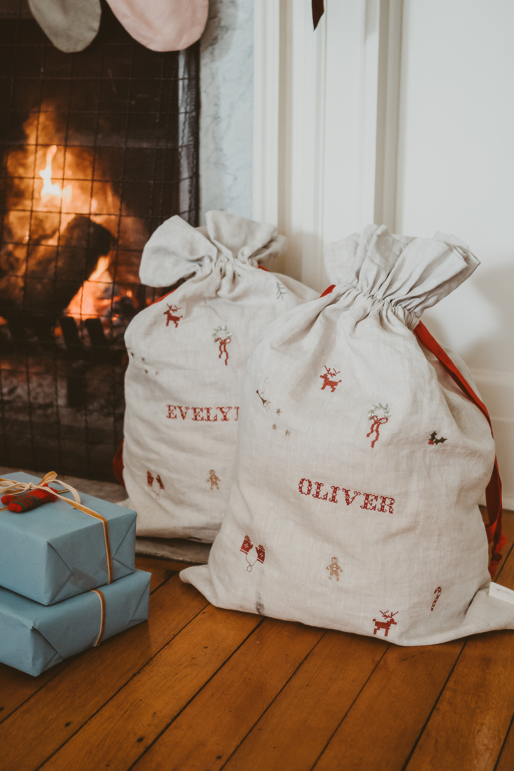 Natural linen Santa sacks with embroidered holiday motifs and personalized names Evelyn and Oliver by fireplace