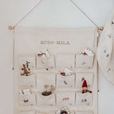 Embroidered Advent Calendar with personalized names, ivory cotton canvas, decorative pockets, and hanging string with tiny bells.