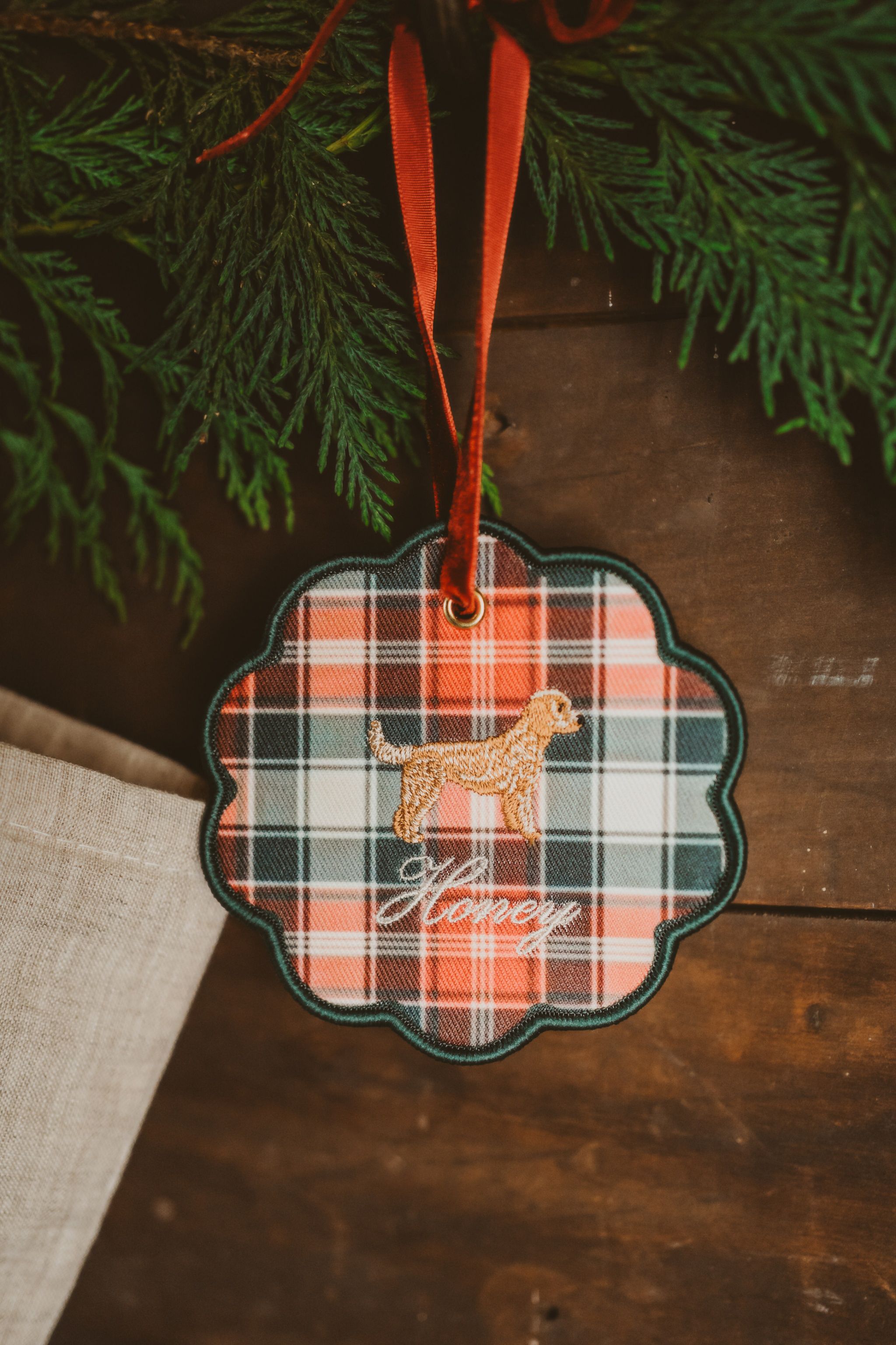 Personalised pet ornament featuring a plaid pattern and embroidered dog design with name "Honey" hanging on a red ribbon in holiday decor setting