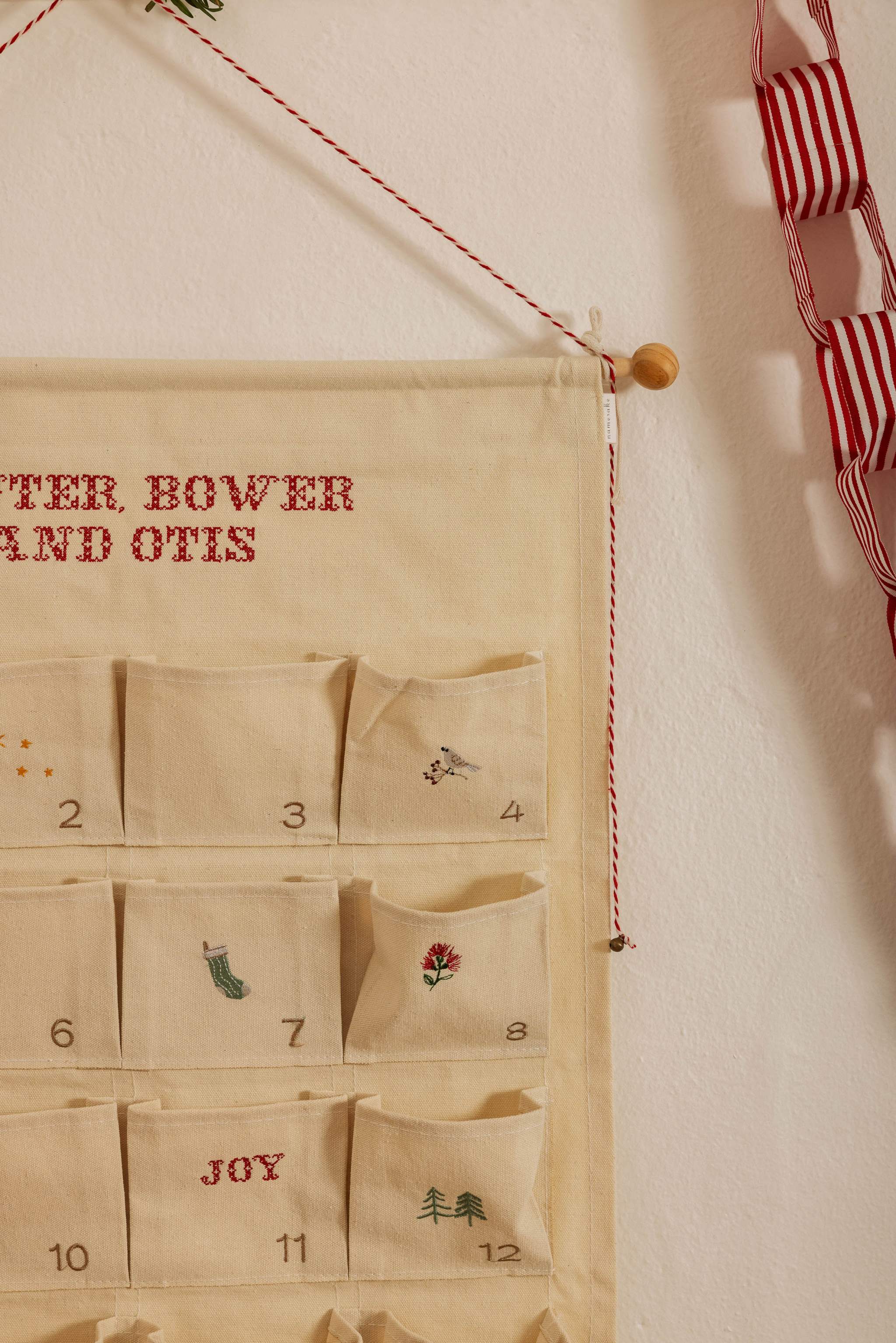 Personalized embroidered advent calendar with Nordic Cross Stitch design on cotton canvas, featuring numbered pockets and hanging wooden dowel.
