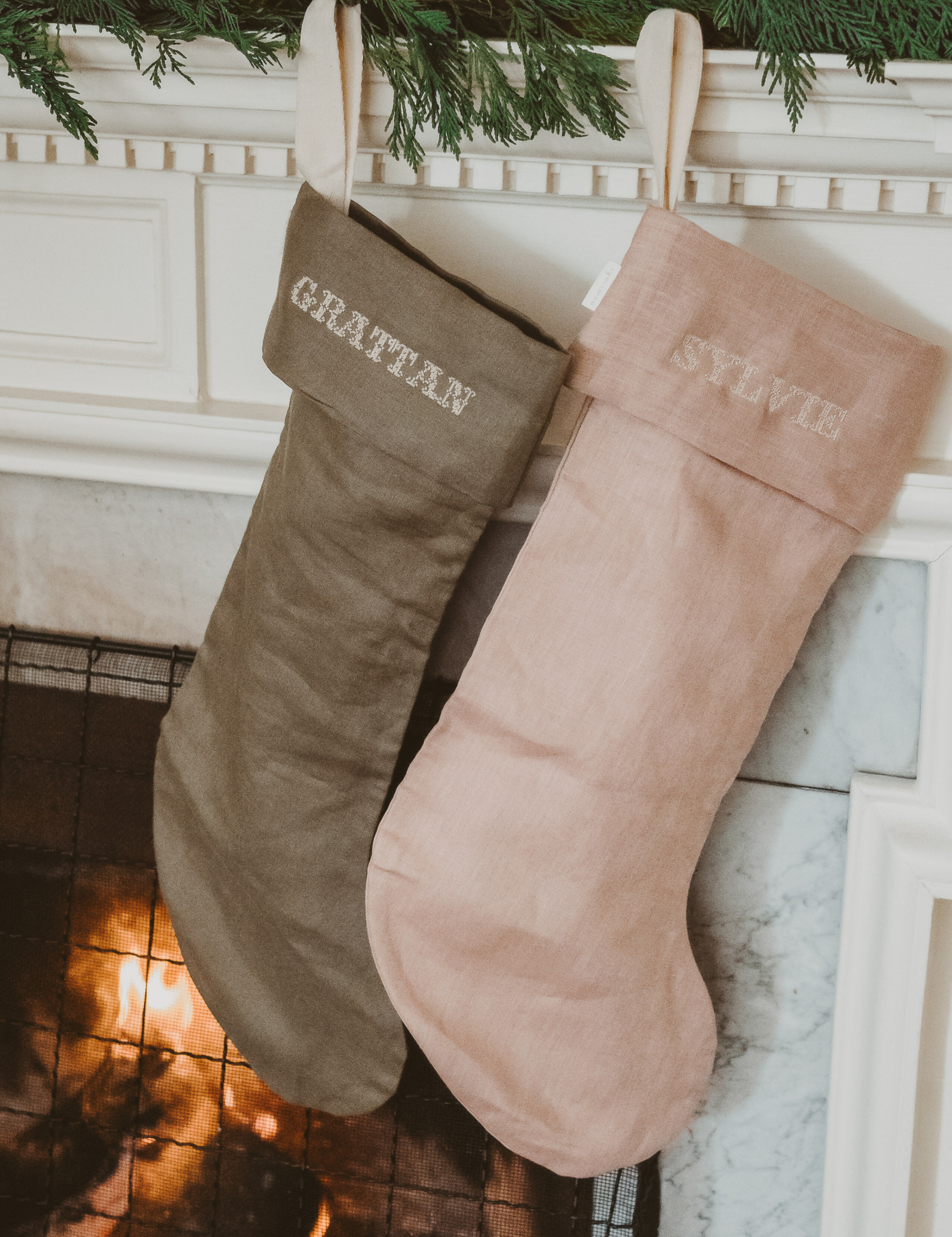 Plain linen stockings in green and pink with embroidered personalized cuffs hanging by a fireplace mantel
