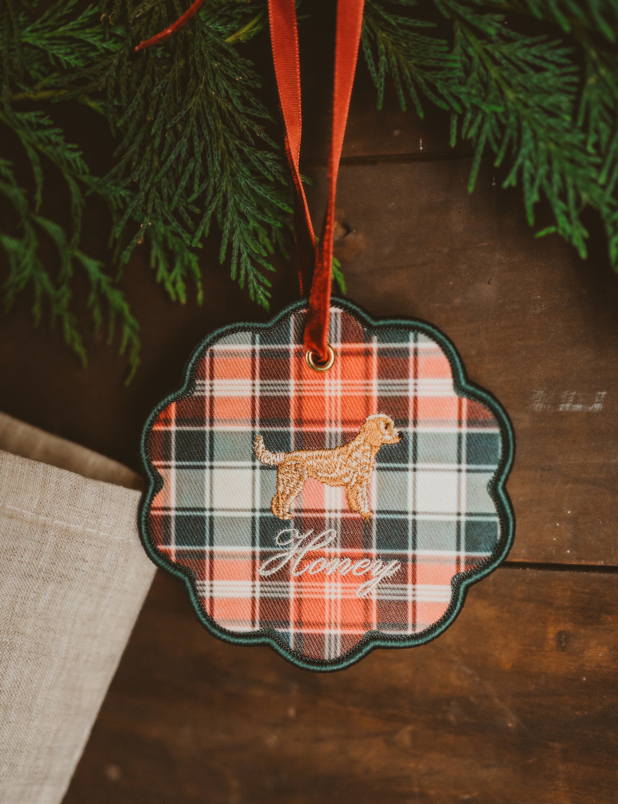 Personalised pet ornament featuring a plaid pattern and embroidered dog design with name "Honey" hanging on a red ribbon in holiday decor setting