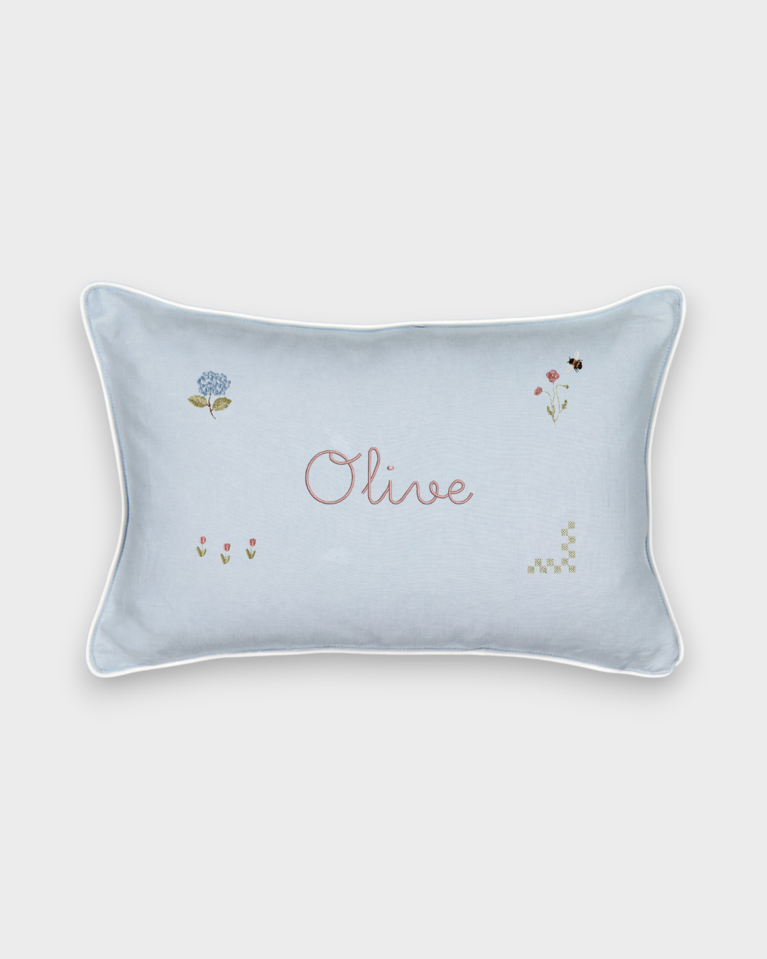 Botanical linen cushion with hand-drawn floral embroidery and personalized name "Olive" on light blue fabric with white piping.